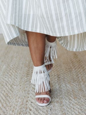 A close-up detail shot of white fringe heeled sandals paired with a flowing striped midi skirt — the kind of shoe that makes a stripe skirt feel like a whole personality.