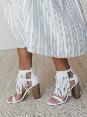 A close-up of white fringe block-heel sandals paired with a striped midi skirt, shot indoors on a natural jute rug — the kind of shoes that make a whole outfit feel more intentional.