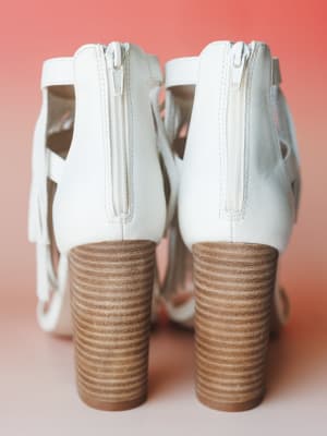 A pair of white leather block-heel sandals shot from behind against a warm coral background, showing off the stacked wood-tone heel and back zip closure with fringe detail peeking through.