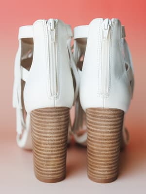 A pair of white leather block-heel sandals shot from behind against a warm coral background, showing off the stacked wood-tone heel and back zip closure with fringe detail peeking through.