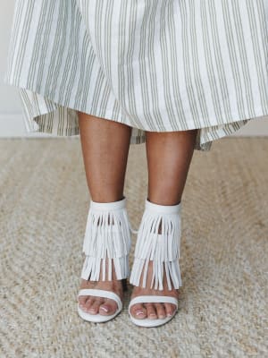 A close-up detail shot of white fringe heeled sandals paired with a flowy olive-striped skirt — the kind of shoe that quietly makes the whole outfit.