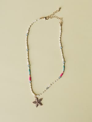 A delicate beaded necklace laid flat on a soft sage background — white seed beads punctuated by pops of hot pink, aqua, and pale blue, anchored by a gold starfish charm that feels like a summer memory you're not ready to let go of.