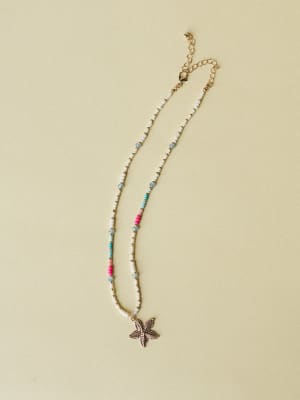 A delicate beaded necklace laid flat on a warm sage background, mixing white seed beads with pops of hot pink, turquoise, and soft blue, finished with a gold starfish pendant and lobster clasp extender chain — total vacation energy.