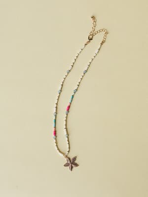 A delicate beaded necklace laid flat on a warm sage background, mixing white seed beads with pops of hot pink, turquoise, and soft blue, finished with a gold starfish pendant and lobster clasp extender chain — total vacation energy.