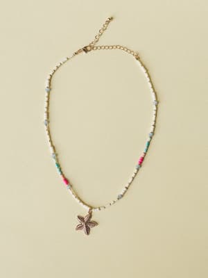 A delicate beaded necklace with white seed beads, scattered pops of pink, aqua, and sky blue, and a textured gold starfish pendant — laid flat on a warm sage background with a gold lobster clasp and extender chain.