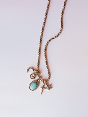 A gold box-chain charm necklace laid flat, layered with celestial and western-inspired pendants — a crescent moon, crystal heart, oval turquoise stone, dagger cross, and pavé star — all with that collected-over-time feel.