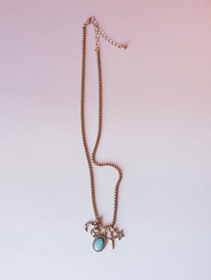 A silver box chain charm necklace laid casually on a soft pink-to-white gradient background, featuring a cluster of mixed charms — turquoise oval stone, crescent moon, crystal heart, small dagger, and star — with an adjustable lobster clasp extender.