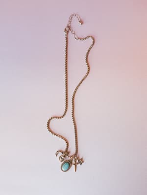 A silver box-chain charm necklace laid against a soft blush gradient background, featuring a cluster of celestial and western-inspired charms — a crescent moon, heart crystal, oval turquoise stone, dagger, and star.