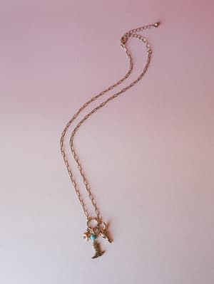 A dainty gold paperclip chain necklace laid casually on a blush pink surface, finished with a cluster of western-inspired charms — a cowboy boot, star, bird, and a small turquoise bead that keeps the whole thing feeling a little sun-worn and well-tra