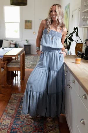 A woman smiles with hands in pockets, wearing the Harlow Wide Leg Chambray Jumpsuit - Blue in a bright kitchen with wooden floors, a colorful rug, and dining furniture in the background.