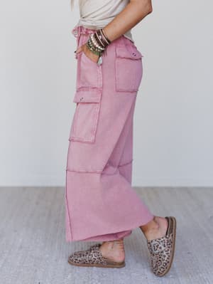Someone stands sideways against a plain background, wearing the Game On Cargo Wide Leg Pant in burgundy with a drawstring stretch waist, a white top, multiple bracelets, leopard print slip-on shoes, and one hand in their pocket.