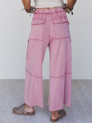 Person stands on a light floor, seen from behind, wearing Game On Cargo Wide Leg Pant in Burgundy with drawstring stretch waist and large back pockets, paired with leopard print slip-on shoes and several bracelets.