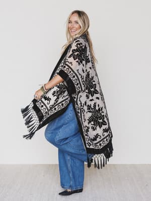 A smiling woman stands against a white background, wearing the Crushin' On You Velvet Kimono - Black over a black top and wide-leg blue jeans. She poses with one leg bent, looking over her shoulder.