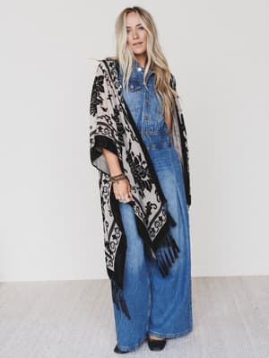 A woman with long blonde hair wears a blue denim jumpsuit and the Crushin' On You Velvet Kimono in black, featuring a boho pattern and fringe. She stands against a plain white wall on a light-colored floor.