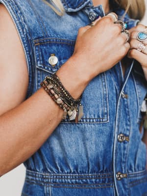 A person wears the Alyda Boho Bracelet Set - Brown, stacked with other boho bracelets and multiple rings, including a turquoise stone ring, their hand raised near their chest.