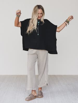 A smiling woman with long blonde hair wears The Nest Moon Glow Texture Knit Top in black, light beige wide-leg pants, and leopard print slip-ons. She raises her arms, accessorized with bracelets and necklaces, posing against a plain background.