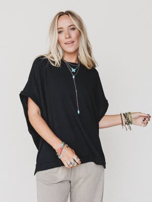 A woman with long blonde hair wears The Nest Moon Glow Texture Knit Top in black and beige pants, accessorized with layered necklaces, bracelets, and rings. She smiles slightly while standing against a plain white background.