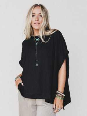 A woman with long blonde hair models The Nest Moon Glow Texture Knit Top in black and beige pants against a white background, styled with layered turquoise necklaces, bracelets, and rings.