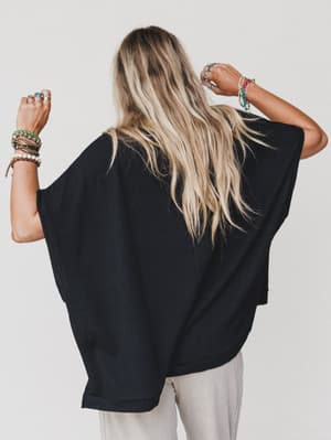 A person with long, wavy blonde hair faces away, wearing The Nest Moon Glow Texture Knit Top - Black and light pants. Their raised arms show multiple colorful bracelets and rings against a plain white background.