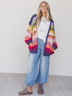 A woman with long blonde hair stands indoors wearing the "Wrapped up in Color Knit Cardigan - Rainbow" over a white tee, loose light blue jeans, and leopard print shoes, accessorized with layered necklaces.