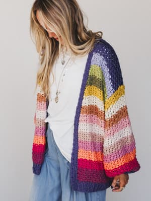 A person with long, blonde hair wears a white shirt, light blue jeans, and the Wrapped up in Color Knit Cardigan – Rainbow, featuring vibrant purple, yellow, pink, orange, and red stripes, while looking down against a plain background.