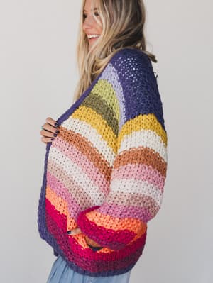 A woman with long blonde hair smiles in profile against a light background while wearing the Wrapped up in Color Knit Cardigan - Rainbow, a chunky boho sweater featuring colorful stripes in purple, yellow, white, pink, orange, and red.