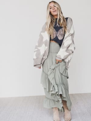 A woman with long blonde hair wears the Wrapped In Stars Oversized Cardigan in taupe, a dark graphic t-shirt, a light green ruffled maxi skirt, and beige boots, standing against a plain white wall and smiling slightly.