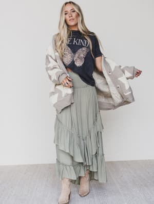 A woman with long blonde hair wears the Be Kind Mineral Washed Graphic Tee - Charcoal, an oversize boho style, paired with a patterned cardigan, sage green ruffled skirt, and beige boots, standing against a plain light background.