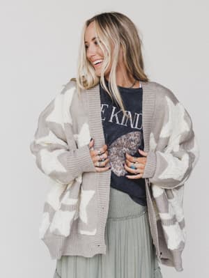 A smiling woman with long blonde hair wears the Wrapped In Stars Oversized Cardigan - Taupe, featuring white star patterns, over a black graphic T-shirt and light green pleated skirt. She completes her look with several rings.