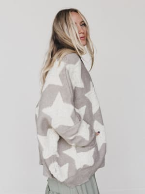 A woman with long blonde hair models the Wrapped In Stars Oversized Cardigan in taupe, featuring bold white stars, as she stands sideways and looks over her shoulder against a plain white background.