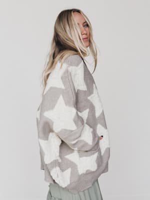 A woman with long blonde hair models the Wrapped In Stars Oversized Cardigan in taupe, featuring bold white stars, as she stands sideways and looks over her shoulder against a plain white background.