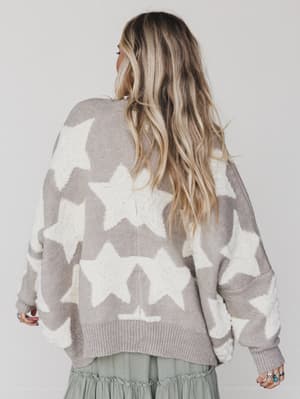 A person with long, wavy blonde hair is seen from behind, wearing the Wrapped In Stars Oversized Cardigan in taupe with large white stars and a green skirt, standing against a plain light background.