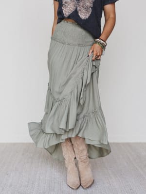 A person in a black T-shirt, layered bracelets, light tan cowboy boots, and the Washed Ashore Ruffled Maxi Skirt – Olive stands on a light-colored carpet for the perfect festival chic look.