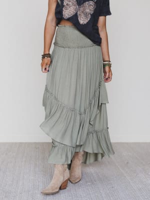 Wearing a dark butterfly-print tee, tan suede ankle boots, and multiple bracelets, a person pairs it all with the Washed Ashore Ruffled Maxi Skirt - Olive on a light woven rug—capturing effortless festival chic style.