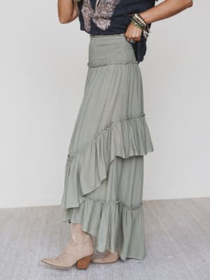 A woman models the Washed Ashore Ruffled Maxi Skirt in olive with a navy graphic tee, bracelets, and beige ankle boots against a plain white backdrop. Her face is hidden, creating an effortless festival chic look.