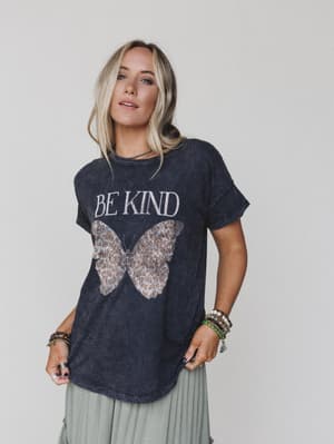 A blonde woman confidently models the Be Kind Mineral Washed Graphic Tee in charcoal, featuring an oversized fit and butterfly graphic, styled with bracelets against a light background.