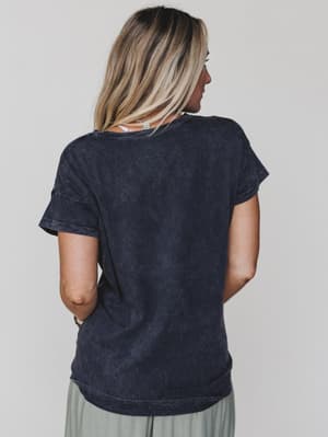 A woman with blonde hair faces away from the camera, wearing the Be Kind Mineral Washed Graphic Tee in charcoal and light gray pants against a plain, light background.