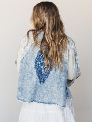 Shot from the back, this acid-washed denim cropped jacket layers over a white tiered skirt — the standout detail is a bold blue embroidered medallion with gold studs centered on the back, with contrast floral and lace panels at the shoulders giving i