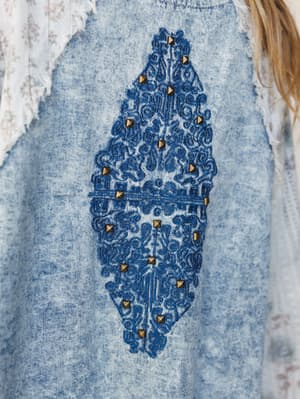 A close-up of an acid wash denim blazer with a striking diamond-shaped embroidered medallion at center front, detailed with brass pyramid studs and fringe trim peeking in at the shoulders — the kind of piece that does the talking for you.