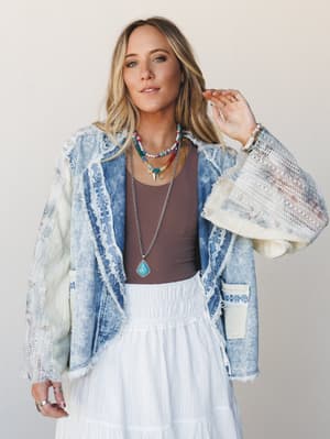 A layered boho look built around an acid-wash denim blazer with lace and crochet trim, styled over a brown tank and white tiered maxi skirt, finished with stacked turquoise and beaded jewelry — easy, expressive, and effortlessly collected.