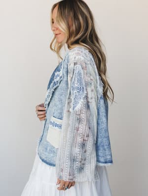 A side-profile shot of a model in a boho denim blazer with mixed-media detailing — frayed edges, embroidery, and lace paneling layered over a white tiered skirt, all shot against a soft neutral studio wall.