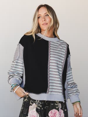 A patchwork mixed-media sweatshirt in black and periwinkle with floral folk-print panels, styled with a dark floral skirt and layered boho bracelets — relaxed and expressive with an artsy, collected feel.