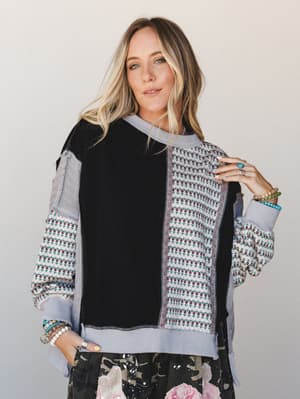 A relaxed patchwork sweatshirt in black and soft lavender with a small floral repeat print — styled with layered turquoise and beaded bracelets and a peek of a dark floral skirt beneath, giving the whole look a collected, lived-in energy.