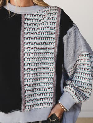 A close-up detail shot of a mixed-fabric pullover top that layers black knit, floral jacquard panels, and a lace sleeve into something that feels like a cozy patchwork project gone exactly right.