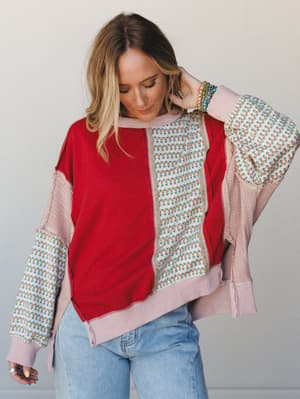 A relaxed mixed-fabric pullover top in patchwork red, blush, and floral print panels worn with light wash jeans — the kind of top that looks intentional without trying too hard.