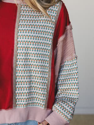 A close-up of a mixed-fabric pullover top that layers a red waffle-knit body with a floral jacquard center panel and patchwork sleeves in blush lace and matching print — the kind of piece that looks like it was always meant to be this way.