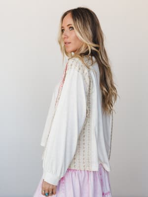 Shot from the side, she's glancing back over her shoulder in a cream top that mixes cozy knit sleeves with a floral-printed woven panel down the back — the kind of top that looks intentional without trying too hard, paired with a soft lavender tiered