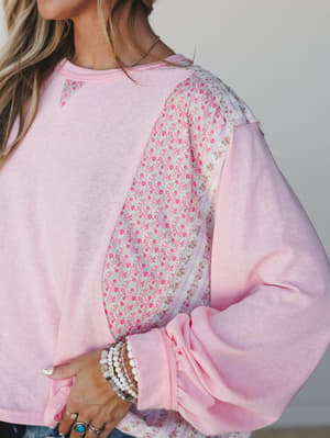 A soft pink sweatshirt top with contrast floral print sleeves and mixed fabric paneling — the kind of piece that feels like your favorite weekend top finally got a little personality.