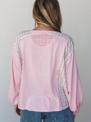 A soft pink long-sleeve top photographed from the back, showing off its sweetest detail — floral patchwork paneling along the shoulders and sides that turns a simple silhouette into something worth a second look.