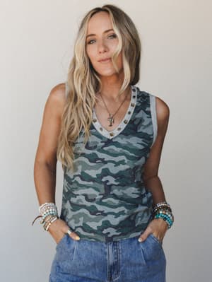 A camo tank top with studded v-neckline and contrast gray trim, styled with stacked boho bracelets and a layered pendant necklace — the kind of effortless, lived-in look that works as hard as she does.